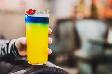 Colorful layered cocktail with blue, green, and yellow hues in a tall glass, garnished with a cherry, held in hand against a blurred background.
