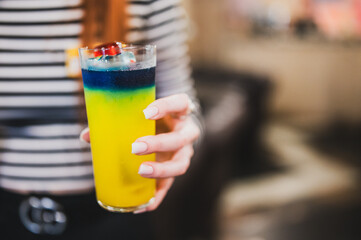 Close-up of a hand holding a layered colorful cocktail in a tall glass, with a summer gradient from...
