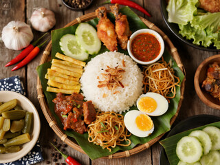 Indonesian coconut rice served with assorted side dishes including fried chicken, tofu, noodles, rendang, boiled egg, and sambal—presented on banana leaves in a rustic woven tray for a hearty and flav