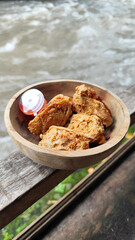 Tahu walik. Crispy browned tofu walik served in a natural wooden bowl with spicy chili sauce, with a blurry river flowing in the background. Menu at an outdoor cafe.
