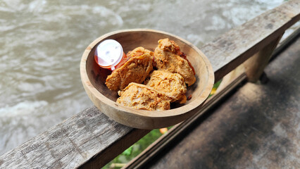 Tahu walik. Crispy browned tofu walik served in a natural wooden bowl with spicy chili sauce, with a blurry river flowing in the background. Menu at an outdoor cafe.