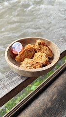 Tahu walik. Crispy browned tofu walik served in a natural wooden bowl with spicy chili sauce, with a blurry river flowing in the background. Menu at an outdoor cafe.