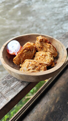 Tahu walik. Crispy browned tofu walik served in a natural wooden bowl with spicy chili sauce, with a blurry river flowing in the background. Menu at an outdoor cafe.