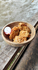 Tahu walik. Crispy browned tofu walik served in a natural wooden bowl with spicy chili sauce, with a blurry river flowing in the background. Menu at an outdoor cafe.