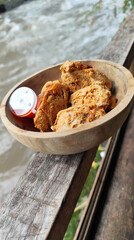 Tahu walik. Crispy browned tofu walik served in a natural wooden bowl with spicy chili sauce, with a blurry river flowing in the background. Menu at an outdoor cafe.