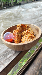 Tahu walik. Crispy browned tofu walik served in a natural wooden bowl with spicy chili sauce, with a blurry river flowing in the background. Menu at an outdoor cafe.