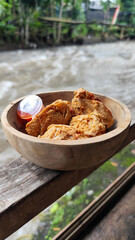 Tahu walik. Crispy browned tofu walik served in a natural wooden bowl with spicy chili sauce, with a blurry river flowing in the background. Menu at an outdoor cafe.