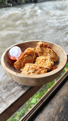 Tahu walik. Crispy browned tofu walik served in a natural wooden bowl with spicy chili sauce, with a blurry river flowing in the background. Menu at an outdoor cafe.