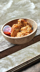 Tahu walik. Crispy browned tofu walik served in a natural wooden bowl with spicy chili sauce, with a blurry river flowing in the background. Menu at an outdoor cafe.