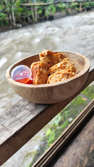 Tahu walik. Crispy browned tofu walik served in a natural wooden bowl with spicy chili sauce, with a blurry river flowing in the background. Menu at an outdoor cafe.