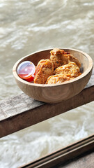 Tahu walik. Crispy browned tofu walik served in a natural wooden bowl with spicy chili sauce, with a blurry river flowing in the background. Menu at an outdoor cafe.