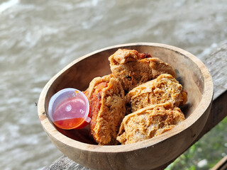 Tahu walik. Crispy browned tofu walik served in a natural wooden bowl with spicy chili sauce, with a blurry river flowing in the background. Menu at an outdoor cafe.
