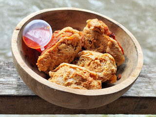 Tahu walik. Crispy browned tofu walik served in a natural wooden bowl with spicy chili sauce, with a blurry river flowing in the background. Menu at an outdoor cafe.