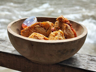 Tahu walik. Crispy browned tofu walik served in a natural wooden bowl with spicy chili sauce, with a blurry river flowing in the background. Menu at an outdoor cafe.