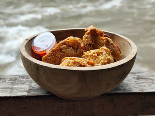 Tahu walik. Crispy browned tofu walik served in a natural wooden bowl with spicy chili sauce, with a blurry river flowing in the background. Menu at an outdoor cafe.