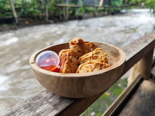 Tahu walik. Crispy browned tofu walik served in a natural wooden bowl with spicy chili sauce, with a blurry river flowing in the background. Menu at an outdoor cafe.