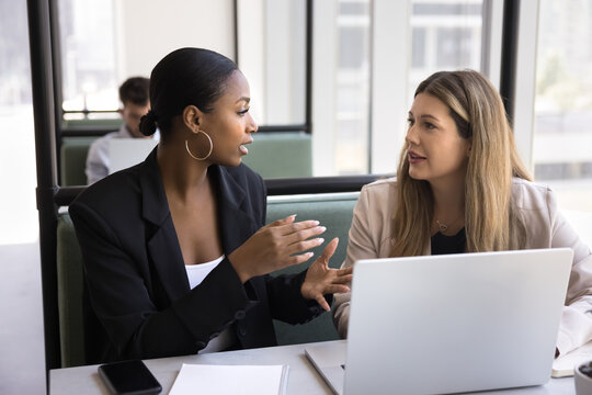 Business conversation in flexible workspace. Mid aged woman client customer focused on discussion with young female legal advisor real estate agent sitting at desk in contemporary coworking hub cabin