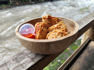 Tahu walik. Crispy browned tofu walik served in a natural wooden bowl with spicy chili sauce, with a blurry river flowing in the background. Menu at an outdoor cafe.