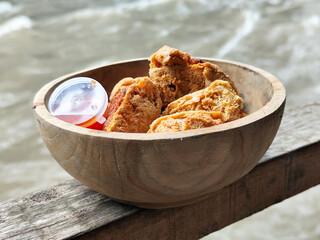 Tahu walik. Crispy browned tofu walik served in a natural wooden bowl with spicy chili sauce, with a blurry river flowing in the background. Menu at an outdoor cafe.