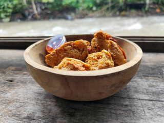Tahu walik. Crispy browned tofu walik served in a natural wooden bowl with spicy chili sauce, with a blurry river flowing in the background. Menu at an outdoor cafe.