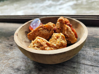 Tahu walik. Crispy browned tofu walik served in a natural wooden bowl with spicy chili sauce, with a blurry river flowing in the background. Menu at an outdoor cafe.