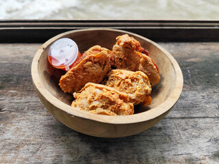 Tahu walik. Crispy browned tofu walik served in a natural wooden bowl with spicy chili sauce, with a blurry river flowing in the background. Menu at an outdoor cafe.