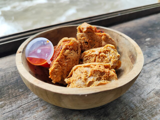 Tahu walik. Crispy browned tofu walik served in a natural wooden bowl with spicy chili sauce, with a blurry river flowing in the background. Menu at an outdoor cafe.