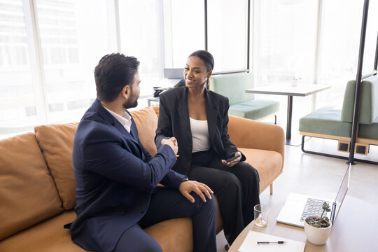 Appreciate for help. Satisfied young businessman in formal suit sit on couch in coworking office shake hand of smiling female consultant advisor lawyer grateful for professional assistance making deal