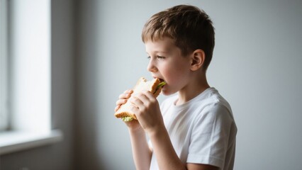 Cute little boy eating sandwich with green salad indoors near window, concept of healthy lifestyle and nutrition, happy child in white t-shirt biting the tasty food during breakfast time
