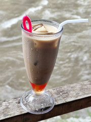 A tall glass of cold chocolate milk with ice cubes and a straw, placed on the edge of a weathered wooden fence with a blurry backdrop of a fast flowing river. Outdoor cafe.