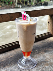 A tall glass of cold chocolate milk with ice cubes and a straw, placed on the edge of a weathered wooden fence with a blurry backdrop of a fast flowing river. Outdoor cafe.