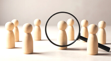 A group of wooden figures with one being highlighted by a magnifying glass on a white background surface