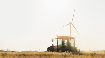 Agricultural tractor is working on the field with wind turbines