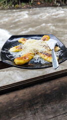 A plate of grilled bananas sprinkled with grated cheese and sweetened condensed milk on a black plate, with a backdrop of a rocky river and blurry green trees. Outdoor cafe.