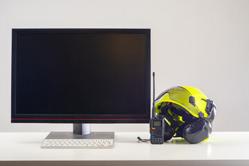 Hardhat with headphones near computer. Worker helmet and walkie-talkie on builder desk. Hardhat with headphones for working in noisy factory. Helmet near black monitor. Hardhat with glasses for eye