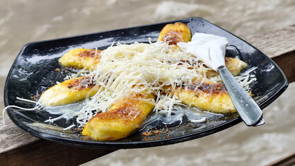 A plate of grilled bananas sprinkled with grated cheese and sweetened condensed milk on a black plate, with a backdrop of a rocky river and blurry green trees. Outdoor cafe.