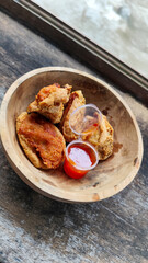 Tahu walik. Crispy browned tofu walik served in a natural wooden bowl with spicy chili sauce, with a blurry river flowing in the background. Menu at an outdoor cafe.