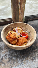 Tahu walik. Crispy browned tofu walik served in a natural wooden bowl with spicy chili sauce, with a blurry river flowing in the background. Menu at an outdoor cafe.