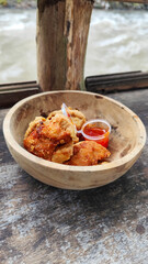 Tahu walik. Crispy browned tofu walik served in a natural wooden bowl with spicy chili sauce, with a blurry river flowing in the background. Menu at an outdoor cafe.