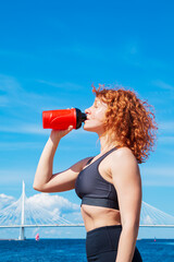Energetic red-haired curly young woman in sportswear drinking water.