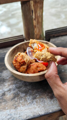 Tahu walik. Crispy browned tofu walik served in a natural wooden bowl with spicy chili sauce, with a blurry river flowing in the background. Menu at an outdoor cafe.