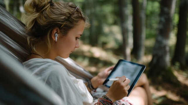 Woman is relaxing in hammock in forest, using tablet and stylus to create or take notes. She appears focused and content, enjoying serene natural surroundings