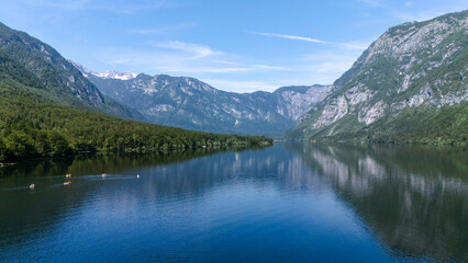 Drone photo of Lake Bohinj in Triglav National Park. Clear blue water mirrors the forest and the Julian Alps while kayaks cross the surface. Taken on a bright summer day. 