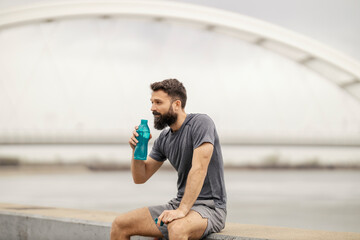 Tired thirsty sportsman sitting on dock and refreshing himself with bottle of water.