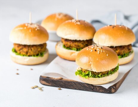 Small vegan lentil burgers with fresh green lettuce and sesame seed buns are neatly arranged on a wooden board on a light table, ready to be enjoyed.