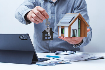 A model home with solar panels and keys in the hands of a realtor. Concepts of home selling and...