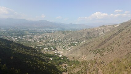 High-angle view of a serene mountain valley town, nestled amongst arid hills and distant mountains under a partly cloudy sky.