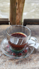 A cup of hot black coffee in a glass cup with matching saucer and spoon, placed on the edge of a weathered wooden fence with a backdrop of a flowing river and blurry green trees. Outdoor cafe menu.
