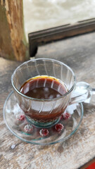 A cup of hot black coffee in a glass cup with matching saucer and spoon, placed on the edge of a weathered wooden fence with a backdrop of a flowing river and blurry green trees. Outdoor cafe menu.