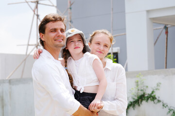 Father and mother are hugging their smiling daughter as they stand in front of the house.